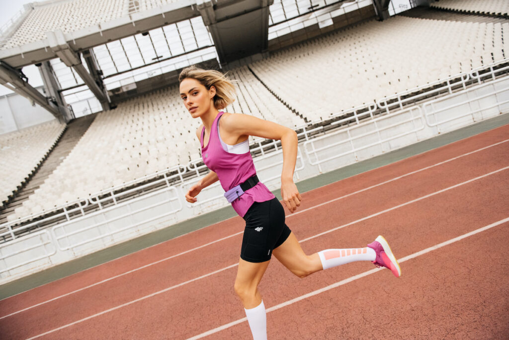 Zdjęcie ze stadionu lekkoatletycznego. W środkowej części widzimy biegnącą kobietę. Ubrana jest ona w różową koszulkę od 4F, czarne spodenki, białe skarpety kompresyjne i czerwone buty dla biegaczy