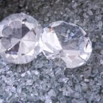 Alrosa to największa spółka zajmująca się produkcją diamentów. Na zdjęciu znajdują się dwa duże, obrobione diamenty leżące na tle dużej ilości mniejszych, nieobrobionych kruszców.