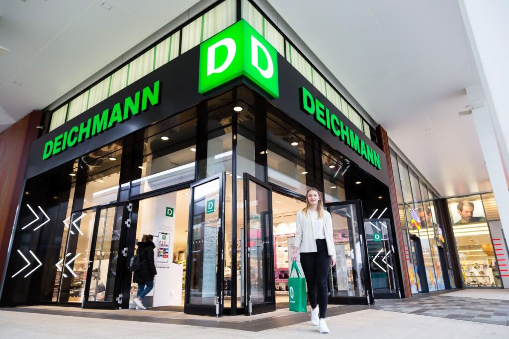 Witryna sklepu Deichmann znajdująca się w centrum handlowym. Na pierwszym planie znajdują się przeszklone drzwi. Nad nimi zielone logo firmy wraz z jej nazwą