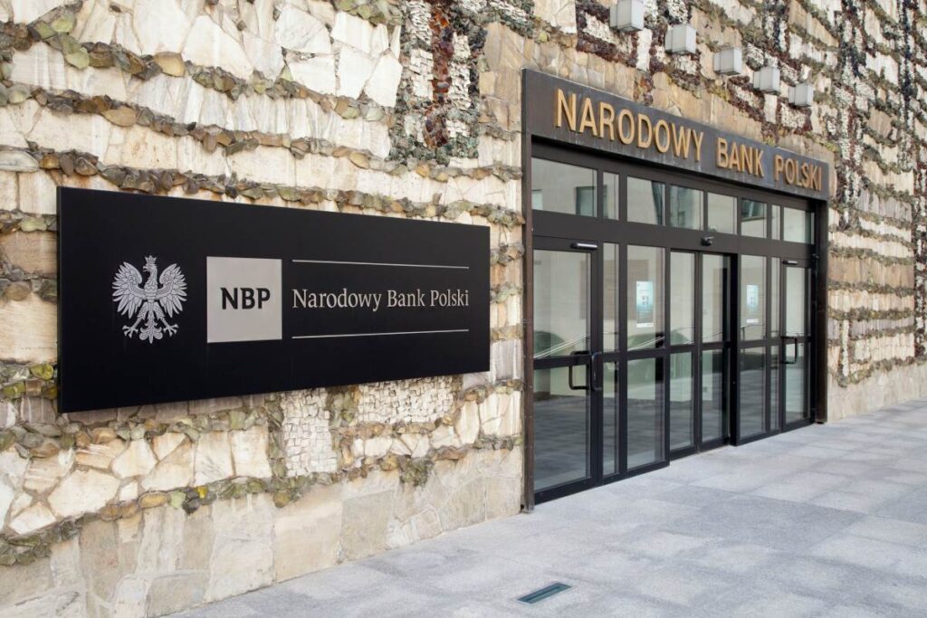 Wejście do budynku Narodowego Banku Polskiego. Duże przeszklone drzwi, nad którymi znajduje się logo NBP