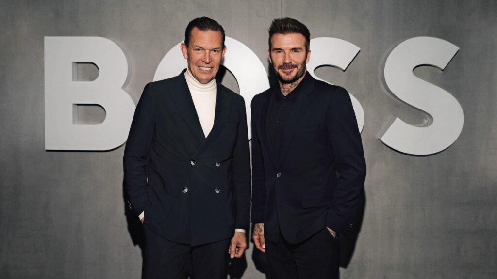 CEO Hugo Boss oraz David Beckam stoją w centralnej części kadru. Są oni ubrani w czarne garnitury. W tle widzimy logo BOSS