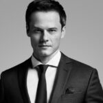 Czarno-białe zdjęcie portretowe. Na pierwszym tle widzimy mężczyznę w białej koszulki, z krawatem oraz ciemną marynarką.