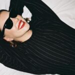 Wartość producenta marki Ray-Ban. Kobieta w czarnym swetrze uśmiecha się leżąc na łóżku. Na twarzy ma czarne okulary marki należącej do EssilorLuxottica