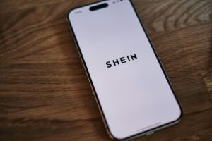 Toksyczne ubrania z Shein. Na zdjęciu widzimy leżący na drewnianym blacie telefon. Na jego ekranie widzimy wyświetlające się czarne logo shein na białym tle.
