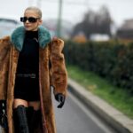 Czy zbyt niska temperatura w pracy może zwolnić nas z wykonywania obowiązków? Na zdjęciu widzimy blondwłosą modelkę idącą wzdłuż ulicy. Kobieta ma na sobie czarne okulary przeciwłoneczne oraz grupy, brązowy kożuch z turkusowym kołnierzem.