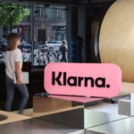 W głównej części zdjęcia znajduje się napis Klarna na różowym tle. Logo znajduje się wewnątrz przestrzeni biurowca.