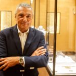 Dyrektor generalny marki Bulgari. Mężczyzna w białej koszuli i w granatowej marynarce stoi z założonymi rękoma. Na jego prawym nadgarstku widnieje zegarek. Mężczyzna znajduje się w salonie podkreślającym jego zawodowy związek z rynkiem dóbr luksusowych.