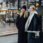 H&M Black Friday 2024. Dwie kobiety blondynki stoją na zaśnieżonej ulicy. Kobieta z lewej ma na sobie długi czarny płaszcz i czarną czapkę. Blondynka z prawej jest ubrana w jasny golf i kożuch. W ręku trzyma czarną torebkę.