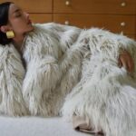 La Mania praca w marketingu. Na zdjęciu kampania La Mania na sezon AW2024. Ciemnowłosa modelka leży na podłodze. Ma na sobie jasne futro, na uszach złote duże kolczyki.