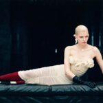 Magda Butrym praca. Sesja z kampanii pre fall 2024. Modelka leży na skórzanej kanapie. Ma na sobie różową sukienkę z motywem róży, czerwone rajstopy, sandały ozdobione różą i czarne skórzane rękawiczki.