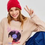 Naoko i Neonail stworzyły wspólną kolekcję bluz wspierających Dziewczynki. Modelka na zdjęciu jest uśmiechnięta i pokazuje znak pokoju. Ma na sobie beżową bluzę z nadrukiem Vintage Baby z kolekcji Naoko i Neonail oraz granatowe spodnie. Na głowie ma czerwoną wełnianą czapkę.