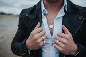 Zbliżenie na klatkę piersiową mężczyzny ubranego w białą koszulę oraz czarną skórę. Na jego szyi znajduje się naszyjnik, natomiast dłonie przyozdabiają sygnety męskie.