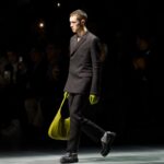 Polski rynek dóbr luksusowych jest częścią światowej branży. Na zdjęciu widzimy modela podczas pokazu domu mody Gucci. Mężczyzna ubrany jest w ciemny garnitur i półbuty. W prawej dłoni trzyma jasną, zieloną torebkę.