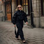 Kobieta ubrana w czarną kurtkę puchową, spodnie bojówki i eleganckie buty spaceruje po miejskim chodniku w zimowej scenerii. W tle widoczne są zabytkowe budynki z drewnianymi drzwiami. Stylizacja przyciąga uwagę minimalistyczną elegancją, idealnie pasując do miejskiego klimatu w dniu, gdy 27 grudnia sklepy są otwarte.