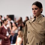 Modelka w klasycznym beżowym trenczu przechadza się po wybiegu, emanując elegancją i pewnością siebie. Jej włosy są starannie zaczesane do tyłu, a wyrazisty makijaż przyciąga uwagę, podkreślając intensywność spojrzenia. W tle rozmyta publiczność z zainteresowaniem obserwuje pokaz, który zachwyca nie tylko stylowymi kreacjami, ale także obietnicą 'biznesowych zaskoczeń 2024'