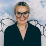Kamila Haciuk, Converse Marketing Managerka CEE. Uśmiechnięta kobieta w okularach o czarnych operawkach, ubrana w czarną koszulę. Kobieta stoi na tle grafiki przedstawiającej rozświetlone przez słońce niebo.