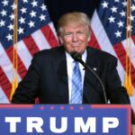 Wyborcze obietnice Trumpa a europejska moda. Donald Trump w czasie wystąpienia. Polityk ubrany w czarną marynarkę, niebieski krawat i białą koszulę. W jego tle znajdują się dwie amerykańskie flagi.