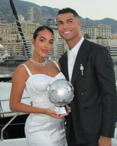 Georgina Rodriguez z męzem, Cristiano Ronaldo na jachcie, z widokiem na panoramę miasta. Georgina ma na sobie białą sukienkę na ramiączkach, kolię na szyi, a włosy są związane na gładko. Obok stoi Cristiano ubrany w garnitur. Georgina trzyma puchar piłkarski.