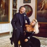 Gianni Versace i jego siotra Donatella Versace pozują razem w zabytkowym, barokowym pomieszczeniu. Gianni ubrany w czarny garnitur patrzy w obiektyw, a siedząca na krześle Donatella, ubrana w aksamitną, czarną suknię wieczorową, patrzy na Gianniego.