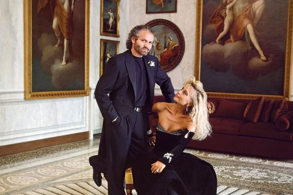 Gianni Versace i jego siotra Donatella Versace pozują razem w zabytkowym, barokowym pomieszczeniu. Gianni ubrany w czarny garnitur patrzy w obiektyw, a siedząca na krześle Donatella, ubrana w aksamitną, czarną suknię wieczorową, patrzy na Gianniego.