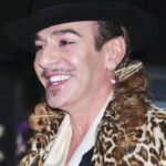 John Galliano z szerokim uśmiechem. Artysta ma na sobie czarny kapelusz oraz płaszcz w jasną panterkę.