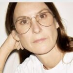 Portret projektantki Louise Trotter. Ma na sobie okulary korekcyjne, rozpuszczone włosy i biały top. Opiera głowę o rękę, pozując do zdjęcia.