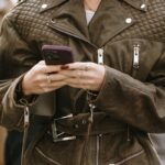 Kobieta ze smartfonem w ręku. Ubrana jest w brązową kurtkę ramoneskę z charakterystycznymi pikowaniami. Idzie ulicą i widać za nią ludzi z dala.