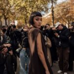 Stylowa kobieta z czarną przestępczością i wyrazistym makijażystem pozuje na tle grupy fotografów. Ma na sobie elegancką, ciemnobrązową kreację z ozdobnymi detalami. Scena, rozgrywająca się wśród jesiennych drzew, przypomina organizację najciekawszych wywiadów 2024