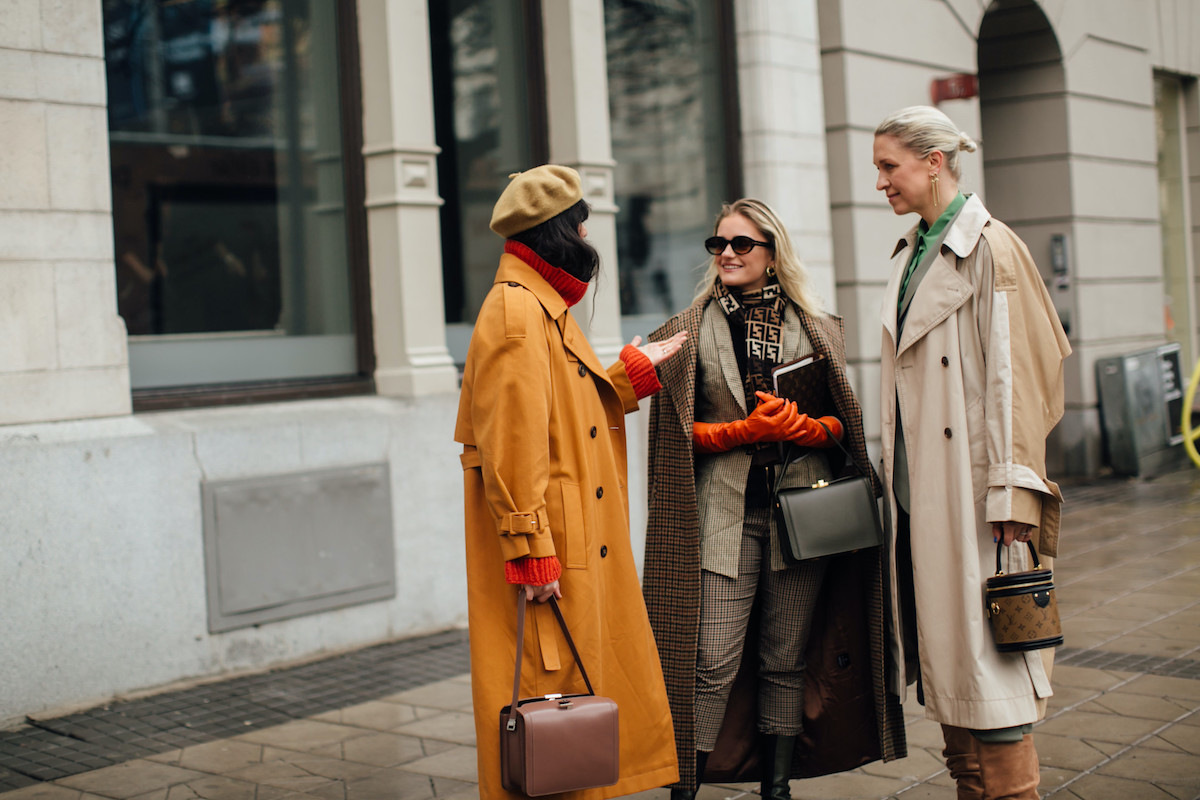 Trzy kobiety w eleganckich płaszczach i akcesoriach w odcieniach beżu, karmelu oraz kratki stoją na ulicy w zimowy dzień i rozmawiają, co kojarzy się z zimowymi zakupami – zwłaszcza że 26 grudnia sklepy otwarte.