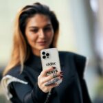 Kobieta w czarnym płaszczu robi sobie selfie smartfonem. Zbliżenie na telefon w białym etui oraz dłoń z czarnymi paznokciami oraz wyrazistymi pierścionkami w kolorze srebrnym.