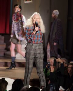 Vivienne Westwood przemawiająca w trakcie pokazu mody do publiczności. Ubrana jest w kraciastą koszulę oraz spodnie w kratkę. Ma rozpuszczone włosy i okulary na nosie.