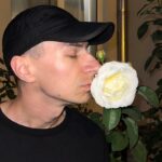 Zbliżenie na twarz mężczyzny, założyciela Pinoko Models. Wojciech Tubaja w czarnej koszulce oraz czarnej czapce z daszkiem wącha białą róże w ogrodzie przed domem.