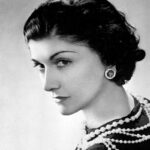 Coco Chanel na starej fotografii. Ma ułożone włosy w fale w stylu retro, ubrana jest w czarną sukienkę oraz sznur pereł. Siedzi tyłem do obiektywu, ale odwraca się do kamery i patrzy prosto na obserwatora.