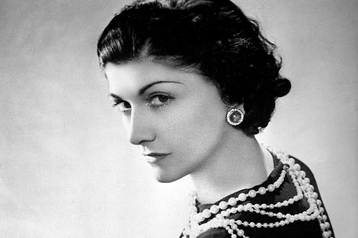 Coco Chanel na starej fotografii. Ma ułożone włosy w fale w stylu retro, ubrana jest w czarną sukienkę oraz sznur pereł. Siedzi tyłem do obiektywu, ale odwraca się do kamery i patrzy prosto na obserwatora.