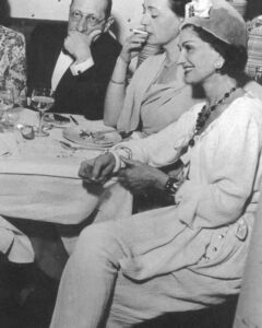 Coco Chanel na starej, czarno-białej fotografii. Ubrana w jasny komplet z marynarką i spodniami. Na głowie ma kapelusz z kokardą. W uszach kolczyki perłowe, a na szyi czarne korale. Siedzi przy stole z innymi gośćmi i szeroko się uśmiecha.