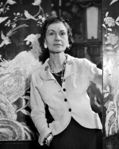 Coco Chanel na starej, czarno-białej fotografii. Ubrana elegancki, jasny żakiet dopasowany do sylwetki oraz małą czarną. Na szyi ma naszyjnik z perłami, w uszach kolczyki z perłami, a na nadgarstkach bransoletki. Znajduje się w pokoju z elegancką tapetą z motywem roślin i ptaków.