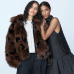 H&M wyprzedaż zimowa 2024. Na zdjęciu kampania H&M holiday na zimę 2024. Dwie modelki przytulone do siebie, mają na sobie nowe projekty marki. Modelka po lewej ma brązowe futerko na zamek, białą koszulę zapiętą pod szyję i szare spodnie materiałowe. Kobieta po prawej ma czarną sukienkę mini bez rękawów, ze zdobieniami przy szyi.
