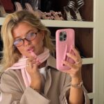 Matilda Djerf, założycielka marki Djerf Avenue, z poważnymi oskarżeniami. Afera, zarzuty. Na zdjęciu skandynawska influencerka z bujnymi blond włosami, przegląda się w przednim aparacie iphona, malując usta różowym błyszczykiem. Dziewczyna ma na sobie okulary, na szyi zawiązany różowy szaliczek oraz beżowy płaszcz. Stoi na tle półki z butami damskimi.