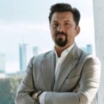 Na zdjęciu znajduje się Dariusz Miłek, prezes CCC. Mężczyzna ubrany w białą koszulę oraz beżową marynarkę, posiadający ciemne włosy i przystrzyżoną brodę.