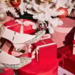 Eleganckie srebrne buty ozdobione kryształkami, zaprezentowane w świątecznym otoczeniu pełnym czerwonych i różowych pudełek na prezenty świąteczne, pod białą choinką ozdobioną bombkami.