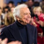 Ralph Lauren - amerykański projektant mody. Życiorys Ralpha Laurena to pasjonująca historia o ciężkiej pracy i spełnianiu marzeń. Na zdjęciu uśmiechnięty projektant podczas swojego pokazu, ubrany w granatowy golf i granatową marynarkę.