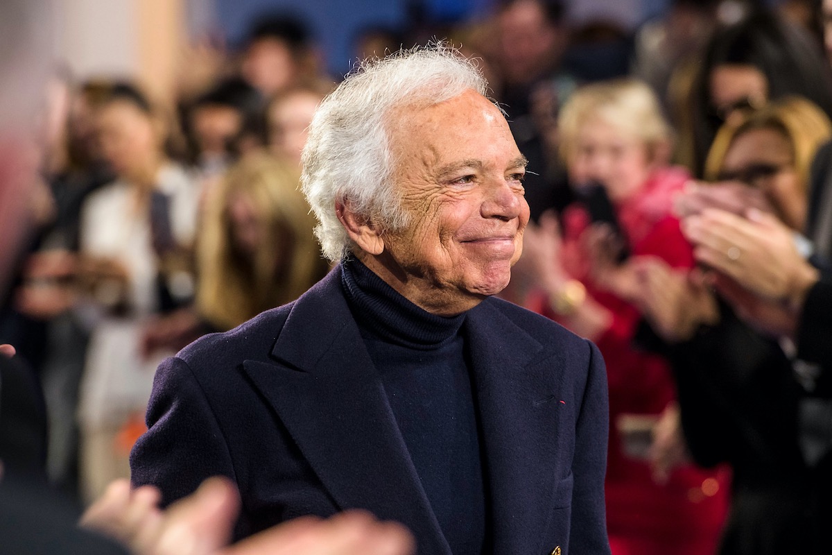 Ralph Lauren - amerykański projektant mody. Życiorys Ralpha Laurena to pasjonująca historia o ciężkiej pracy i spełnianiu marzeń. Na zdjęciu uśmiechnięty projektant podczas swojego pokazu, ubrany w granatowy golf i granatową marynarkę.