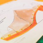 Na zdjęciu widoczny jest projekt obuwia, przedstawiony na papierze w formie kolorowego szkicu technicznego. Rysunek szczegółowo ukazuje konstrukcję buta, w tym jego profil, kształt obcasa oraz detale, takie jak nity czy przeszycia. Projekt został starannie wykonany, co sugeruje profesjonalne podejście do wzornictwa przemysłowego. Fraza „bankructwo fabryki w Pile” może odnosić się do potencjalnych trudności produkcyjnych lub upadku zakładu, w którym realizowano podobne projekty, co wpłynęło na przerwanie takich procesów kreatywnych.