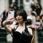 Zdjęcie przedstawia stylową kobietę w czarnej sukience, robiącą selfie smartfonem na tle tłumu. Jej pewny siebie wygląd symbolizuje wpływ mediów społecznościowych na modę i styl życia. W kontekście zakazu TikToka w USA, obraz ukazuje znaczenie platformy dla autopromocji i kreowania trendów w modzie.