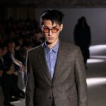 Elegancko ubrany model w okularach przeciwsłonecznych prezentuje stylowy garnitur na tle tłumu podczas pokazu mody. Zdjęcie podkreśla profesjonalizm i atmosferę prestiżu, które mogą kojarzyć się z tematem 'ranking perspektyw 2025', symbolizując sukces i aspiracje.