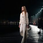 Zdjęcie przedstawia modelkę w eleganckiej, jasnej stylizacji, kroczącej po wybiegu podczas wieczornego pokazu Chanel. W tle widać subtelnie oświetloną scenerię, która podkreśla prestiż i luksusowy charakter wydarzenia. Atmosfera pokazu symbolizuje nie tylko kunszt mody, ale także nową erę dla marki, w kontekście ostatnich wydarzeń - zmiany w zarządzie Chanel, które mają wprowadzić świeże spojrzenie na przyszłość ikony luksusu.
