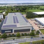 Zdjęcie przedstawia wizualizację nowoczesnego centrum logistycznego Panattoni, zlokalizowanego w Swindon, południowa Anglia. Obiekt posiada rozległy dach pokryty panelami słonecznymi, co podkreśla jego ekologiczne rozwiązania. Na pierwszym planie widoczna jest droga prowadząca do centrum, a w tle otaczająca je zieleń oraz inne budynki przemysłowe i biurowe.