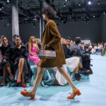 Modelka na wybiegu podczas Fashion Week AW2025, prezentująca brązowy, oversizowy płaszcz zestawiony z pomarańczowymi butami z kokardą oraz torebką w panterkę. Publiczność, ubrana w eleganckie stroje, uważnie obserwuje pokaz w nowoczesnym otoczeniu z pastelową podłogą i industrialnym oświetleniem.