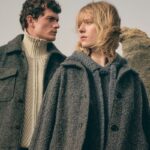 Para modeli ubrana w eleganckie, szare płaszcze i swetry, stojąca na tle surowego krajobrazu. Mężczyzna nosi ciemny, wełniany płaszcz z kołnierzem oraz sweter o wzorze plecionki, natomiast kobieta ma na sobie szary, wełniany płaszcz z guzikami i luźny sweter. Stylizacja w odcieniach szarości oddaje minimalistyczny, nowoczesny charakter mody. W tle widoczne są skały, które dodają surowego, naturalnego klimatu. P&C przejmie Sinn – dwa światy mody, pełne tradycji i nowoczesności, łączą się w jeden.