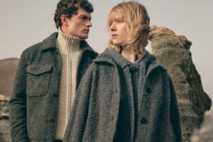 Para modeli ubrana w eleganckie, szare płaszcze i swetry, stojąca na tle surowego krajobrazu. Mężczyzna nosi ciemny, wełniany płaszcz z kołnierzem oraz sweter o wzorze plecionki, natomiast kobieta ma na sobie szary, wełniany płaszcz z guzikami i luźny sweter. Stylizacja w odcieniach szarości oddaje minimalistyczny, nowoczesny charakter mody. W tle widoczne są skały, które dodają surowego, naturalnego klimatu. P&C przejmie Sinn – dwa światy mody, pełne tradycji i nowoczesności, łączą się w jeden.